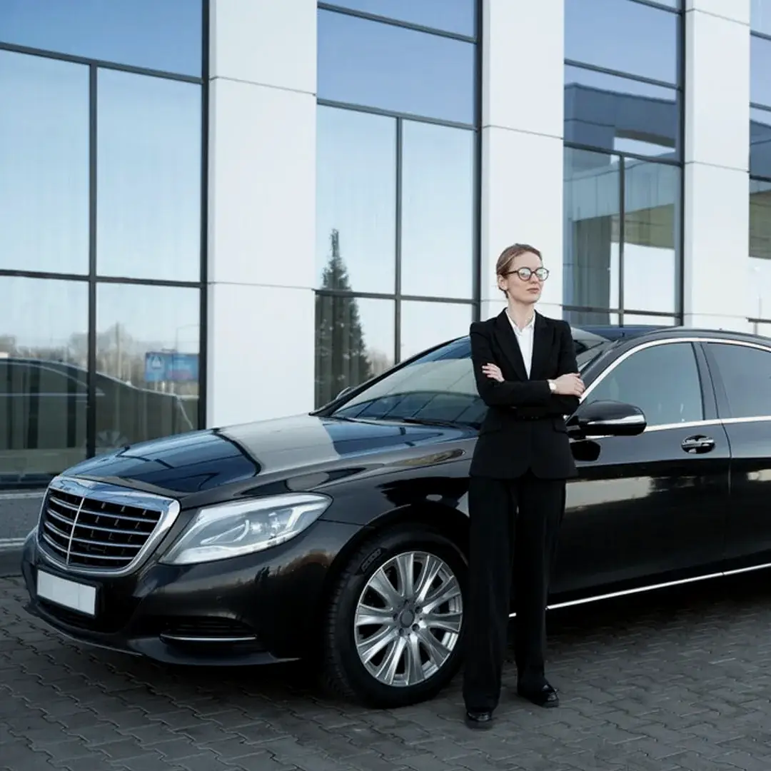 A chauffeur in a black suit waits beside a black sedan outside a glass office building.