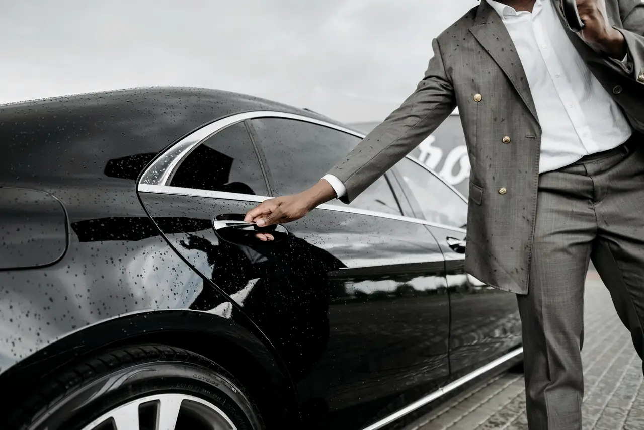 A chauffeur in a grey suit opens the rear door of a black sedan in the rain.